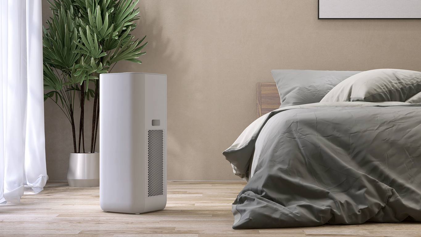 White modern design air purifier, dehumidifier in beige brown wall bedroom, gray cover sheet bed, tropical palm tree in sunlight on wood parquet floor