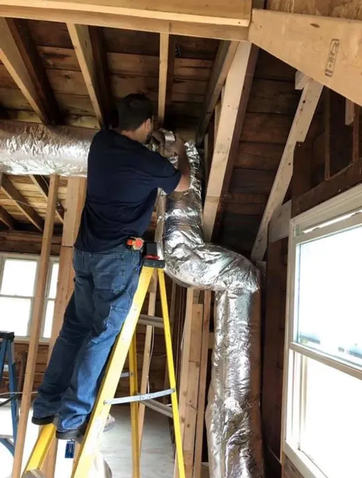 HVAC Blog Man working on duct installation while standing on a ladder inside an unfinished room.